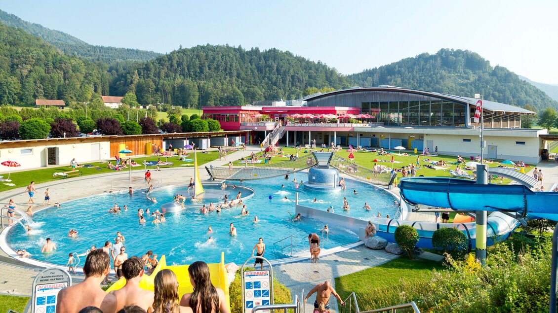 Ein großer Schwimmbereich mit vielen Menschen und einem Rutschenbereich im Freizeitpark Frohnleiten. Im Hintergrund sind grüne Hügel und ein modernes Gebäude zu sehen. | © TV Region Graz - Rene Vidalli