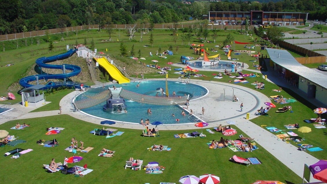 Ein schöner Freizeitpark mit einem großen Schwimmbecken und Wasserrutschen. Viele Menschen entspannen sich auf Liegen im grünen Gras. | © TV Region Graz - Rene Vidalli