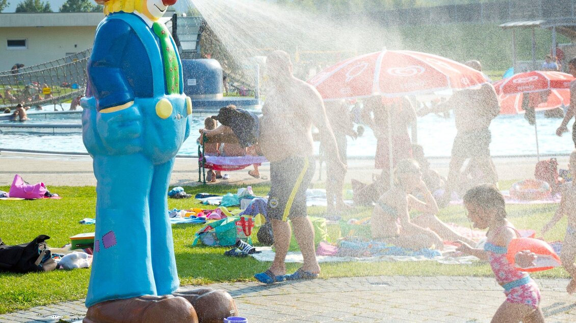 Ein bunter Clown spritzt Wasser in einem Freizeitbad. Im Hintergrund sind Menschen beim Sonnenbaden und Schwimmen zu sehen. | © TV Region Graz - Rene Vidalli