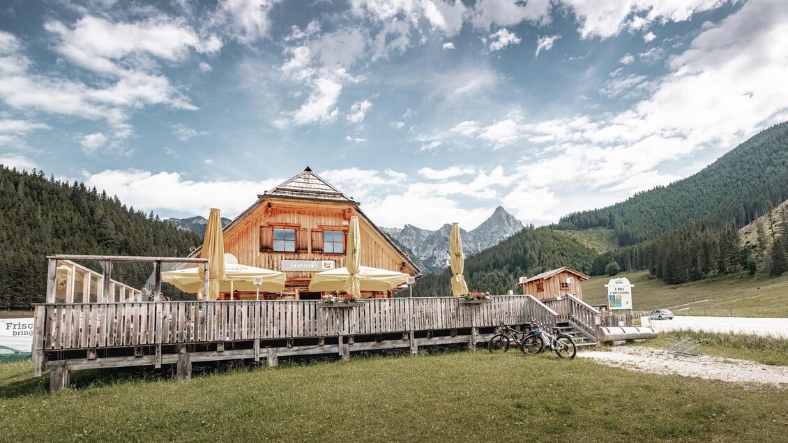 Eine gemütliche Berghütte mit einer Terrasse umgeben von grünen Wiesen und Bergen. Der Himmel ist teilweise bewölkt und verleiht der Szene eine entspannte Atmosphäre. | © Thomas Sattler
