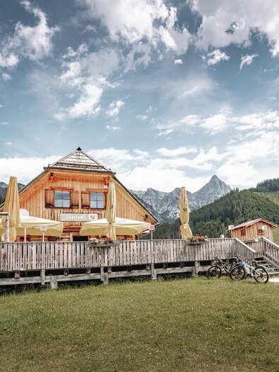 A cozy mountain cabin with a terrace surrounded by green meadows and mountains. The sky is partially cloudy, giving the scene a relaxed atmosphere. | © Thomas Sattler
