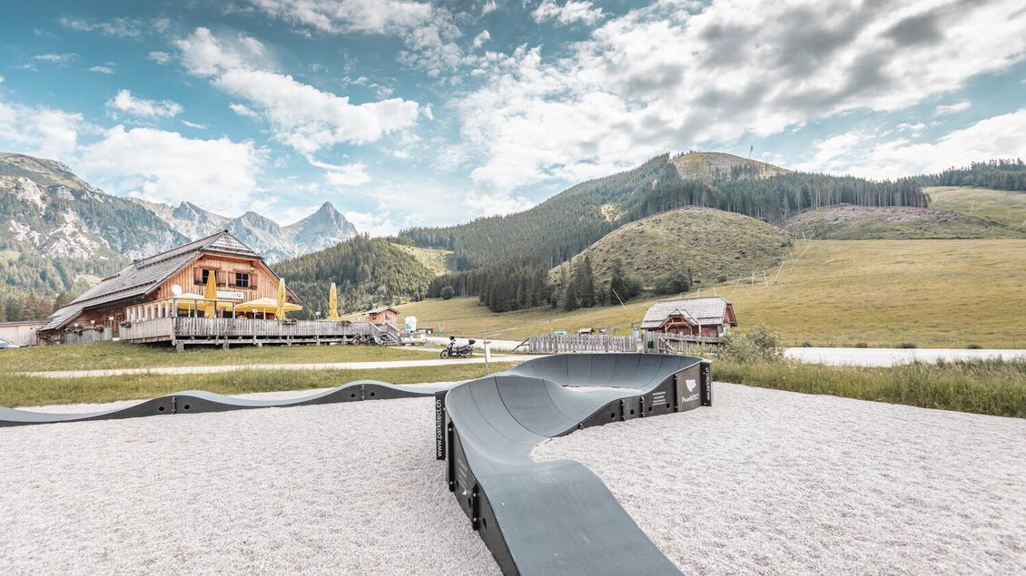 Eine moderne Pumptrack-Strecke in einer bergigen Landschaft. Im Hintergrund sind saftige Wiesen und majestätische Berge zu sehen. | © Thomas Sattler