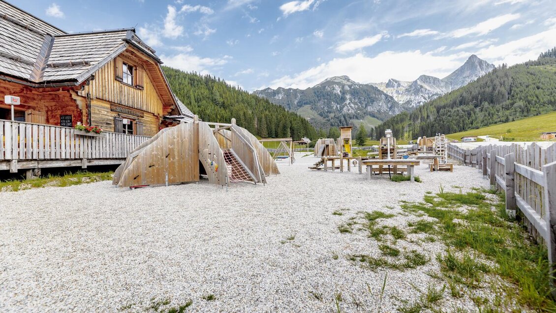 Ein Spielplatz mit Holzstrukturen und einer gemütlichen Berghütte im Hintergrund. Umgeben von grünen Wiesen und Bergen unter einem klaren Himmel. | © Thomas Sattler