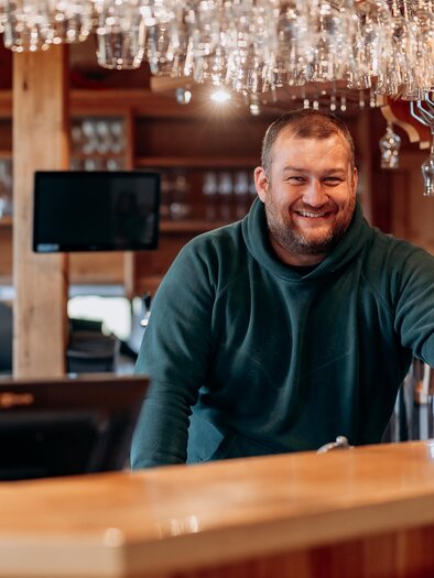 A smiling bartender stands behind the counter holding a wine glass. In the background, glasses hang from the ceiling, and the atmosphere is inviting. | © Stift Admont/Christoph Draxl
