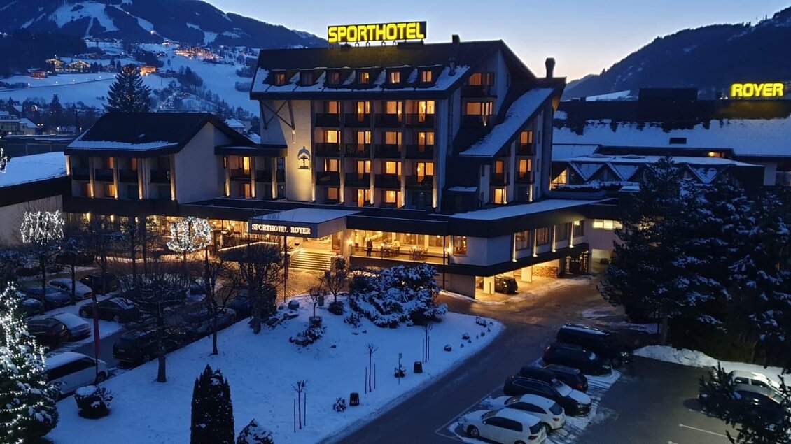 Ein eindrucksvolles Sporthotel im Schnee mit beleuchtetem Eingangsbereich. Die Umgebung ist malerisch mit Bergen im Hintergrund. | © Herbert Raffalt