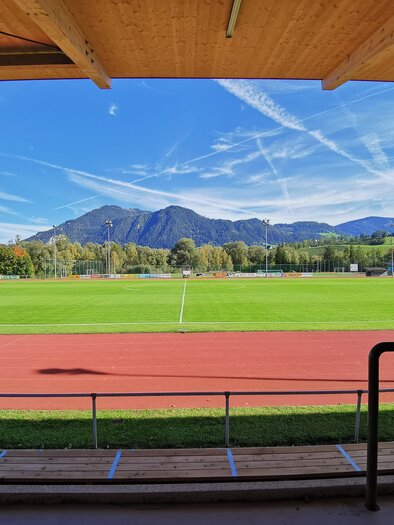 A wide football field with green grass and a red running track. In the background, there are mountains and a clear blue sky. | © Marktgemeinde Stainach-Pürgg