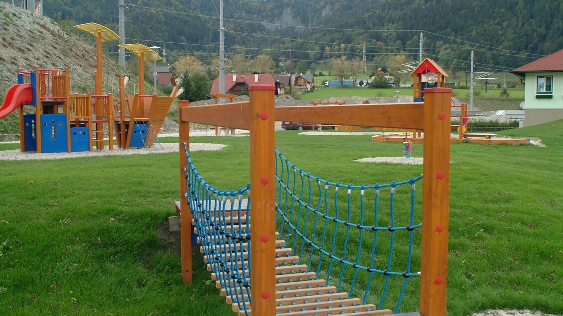Ein Spielplatz mit einer Holzbrücke und Kletterelementen. Im Hintergrund sind Berge und Häuser zu sehen. | © Marktgemeinde Stainach-Pürgg