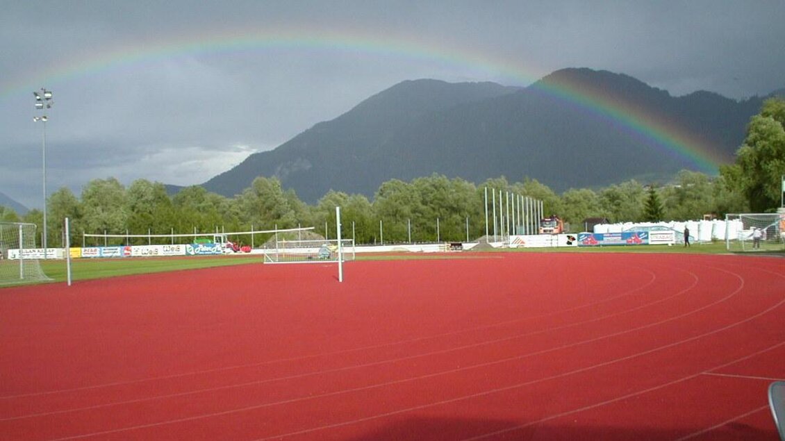 Ein Sportplatz mit rotem Tartanbelag, umgeben von Bäumen und Bergen. Über dem Platz spannt sich ein Regenbogen am grauen Himmel. | © Marktgemeinde Stainach-Pürgg