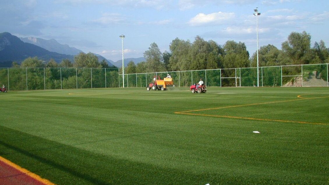 Ein gepflegtes Fußballfeld mit Kunstrasen und grünen Bäumen im Hintergrund. Im Vordergrund sind Traktoren, die das Spielfeld bearbeiten. | © Marktgemeinde Stainach-Pürgg