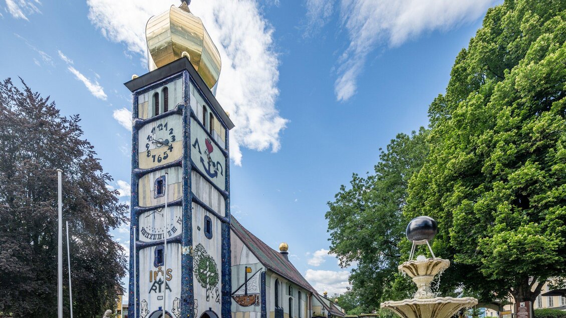 Ein auffälliger Turm mit einer goldenen Kuppel und bunten Wandmalereien. Im Hintergrund sind üppige Bäume und ein blauer Himmel zu sehen. | © TV Region Graz - Die Abbilderei