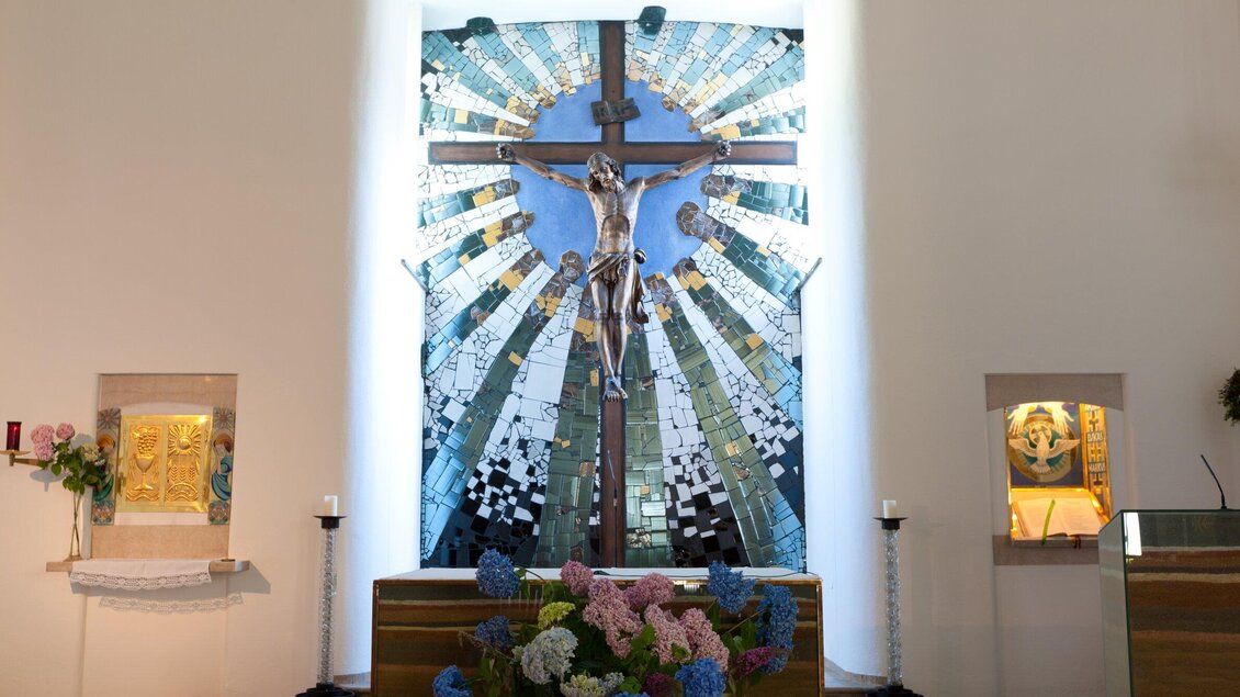 Ein Kreuz mit einem Korpus im Mittelpunkt einer Kirche, umgeben von bunter Glasmalerei. Der Altar ist mit Blumen geschmückt. | © TV Region Graz - Harry Schiffer