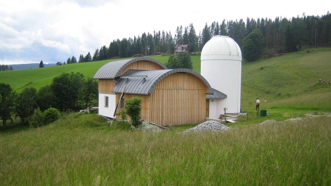 Ein modernes Gebäude mit einer gewölbten Holzfassade und einem kuppelartigen Turm steht in einer grünen Landschaft. Im Hintergrund sind sanfte Hügel und Bäume zu sehen. | © TV Region Graz - Eberhardt