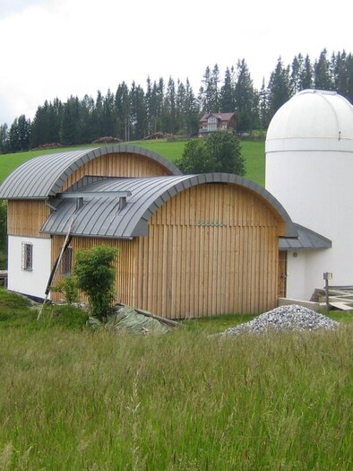 Ein modernes Gebäude mit einer gewölbten Holzfassade und einem kuppelartigen Turm steht in einer grünen Landschaft. Im Hintergrund sind sanfte Hügel und Bäume zu sehen. | © TV Region Graz - Eberhardt