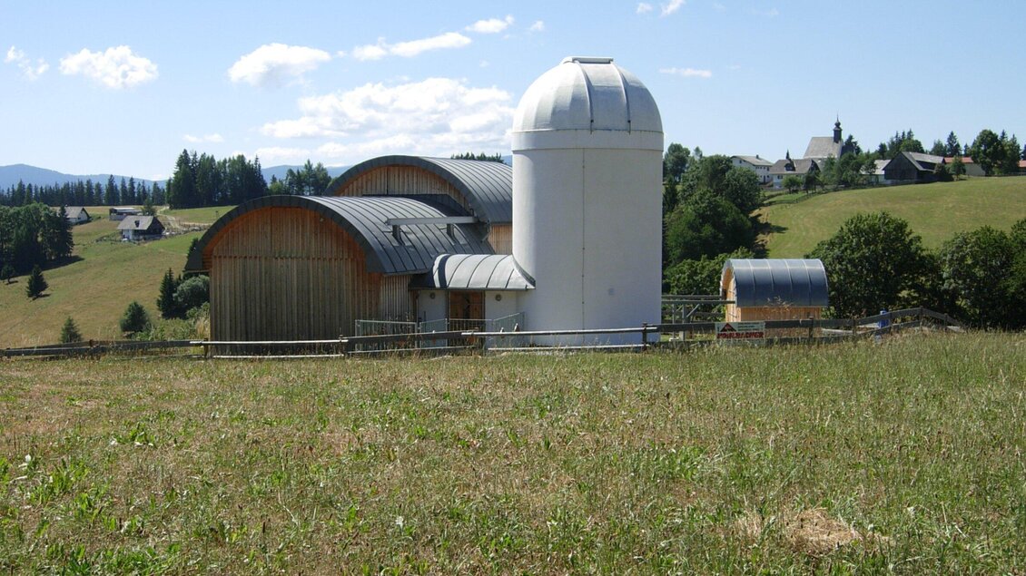 Ein modernes Gebäude mit einer Ziegeldachstruktur und einem runden, weißen Turm steht auf einer Wiese. Im Hintergrund sind hügelige Landschaften und kleine Gebäude zu sehen. | © TV Region Graz - Gerald Lasnik