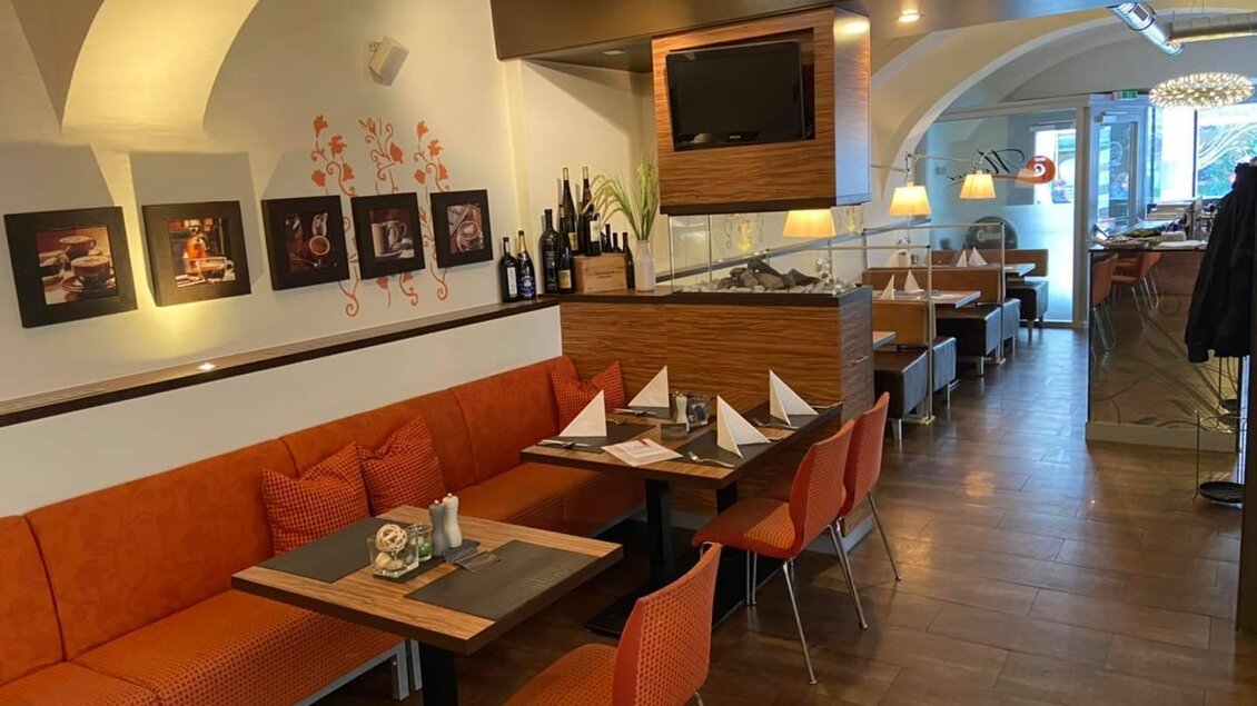 Ein gemütliches Restaurant mit orangefarbenen Sitzen und eleganten Tischen. Kunstwerke schmücken die Wände und eine einladende Atmosphäre vermittelt ein modernes Ambiente.