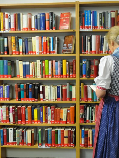 Eine Frau in traditioneller Kleidung steht vor einem Bücherregal. Im Hintergrund sind viele farbige Bücher sichtbar. | © Stadtgemeinde Bad Aussee