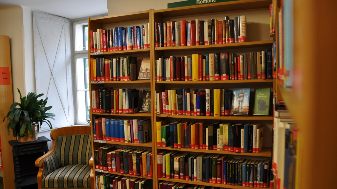 Ein Bücherregal voller Romane in einer Bibliothek. Neben dem Regal steht ein bequemer Sessel mit Kissen. | © Stadtgemeinde Bad Aussee