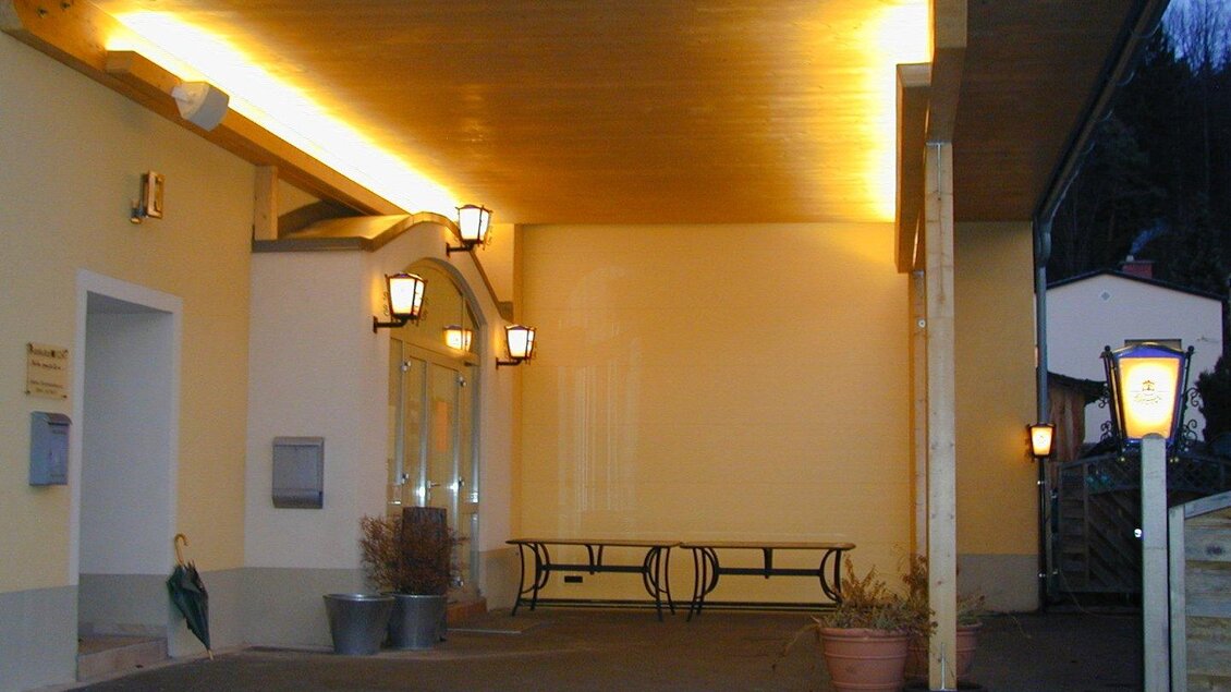 Ein einladender Eingangsbereich mit beleuchtetem Vordach und einer Bank. Die Wände sind hell gestrichen, und es gibt einige Pflanzgefäße. | © Stadtheuriger Trofaiach