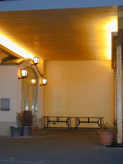 Ein einladender Eingangsbereich mit beleuchtetem Vordach und einer Bank. Die Wände sind hell gestrichen, und es gibt einige Pflanzgefäße. | © Stadtheuriger Trofaiach