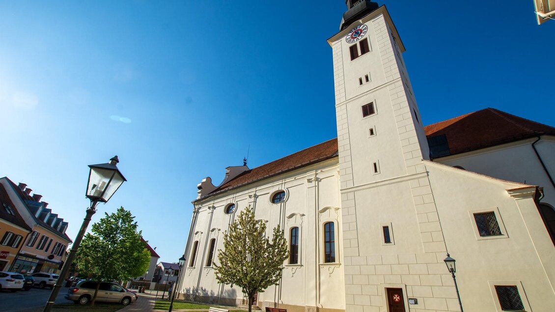 Eine elegante Kirche mit einem hohen Turm und klar blauem Himmel. Auf dem Platz sind einige Bäume und Laternen zu sehen. | © Pfarrverband Fürstenfeld