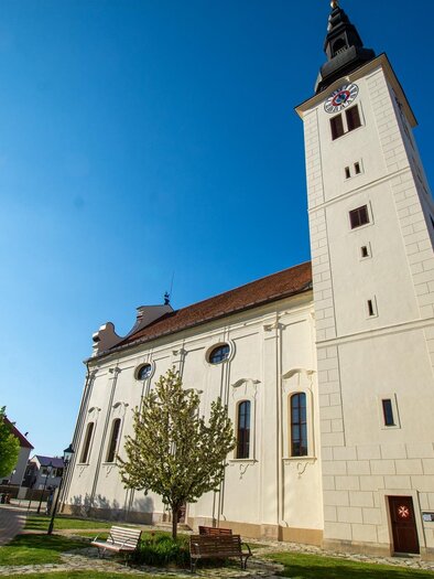 Eine elegante Kirche mit einem hohen Turm und klar blauem Himmel. Auf dem Platz sind einige Bäume und Laternen zu sehen. | © Pfarrverband Fürstenfeld