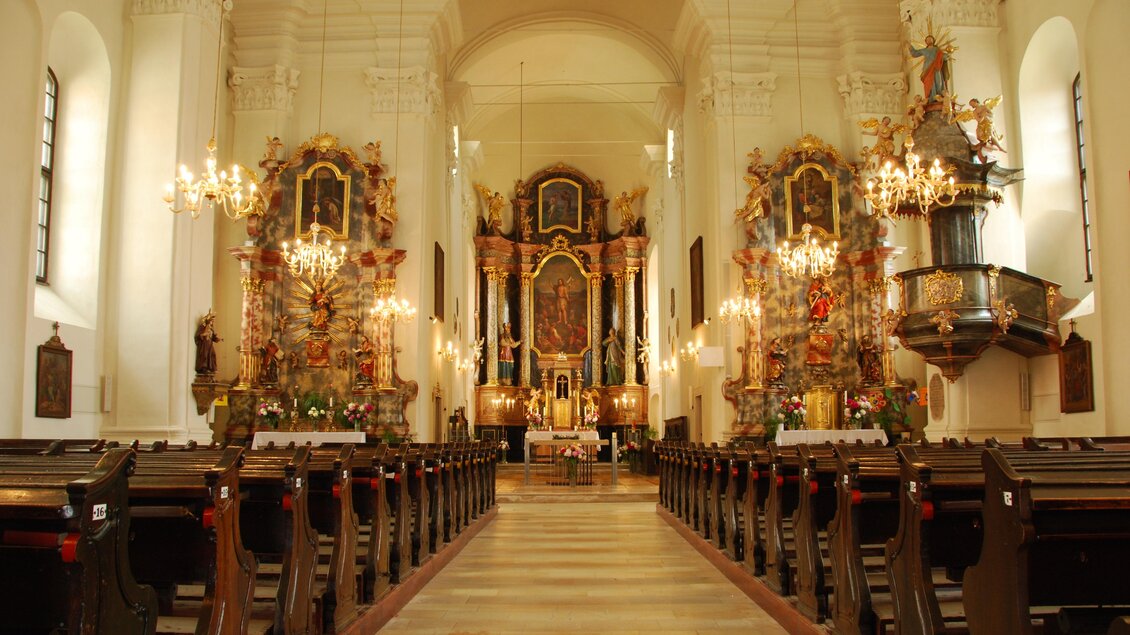 Ein wunderschöner Kircheninnenraum mit hohen Decken und prächtigen Altären. Reihen von Holzbänken führen zum Altar, der festlich dekoriert ist.