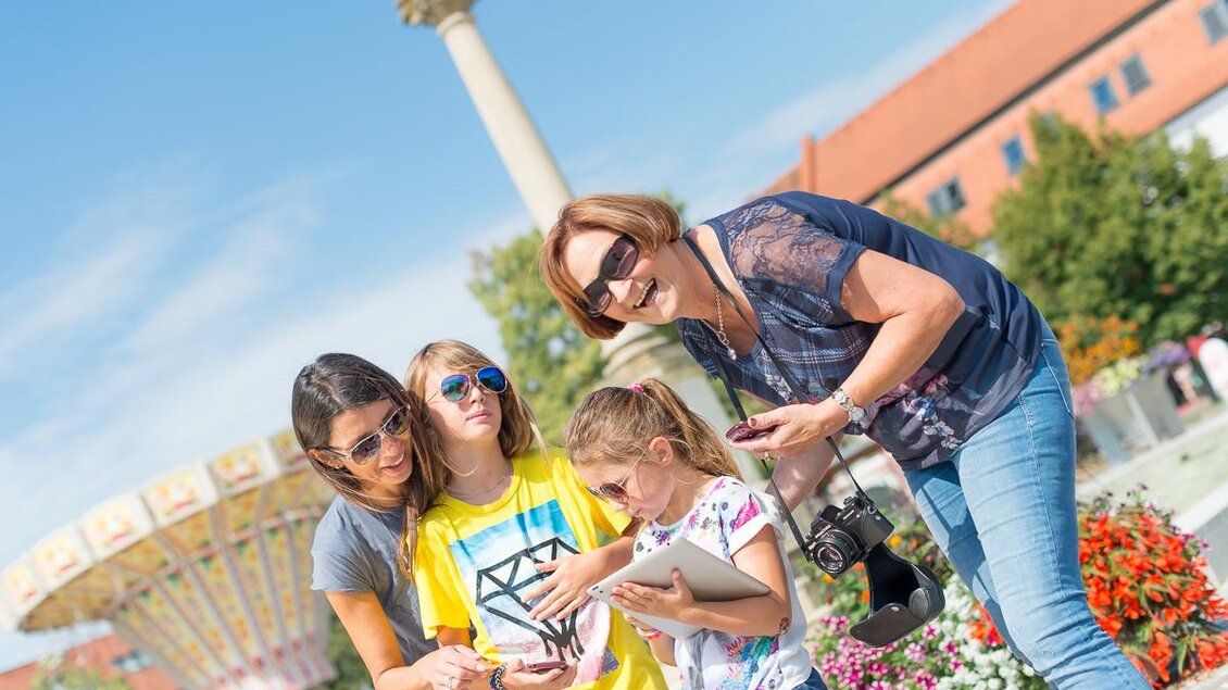 Eine Gruppe von Frauen und Kindern genießt einen sonnigen Tag im Freien. Sie schauen zusammen auf ein Handy und lachen, umgeben von Blumen und einem Vergnügungspark. | © Tourismusverband Thermen- Vulkanland
