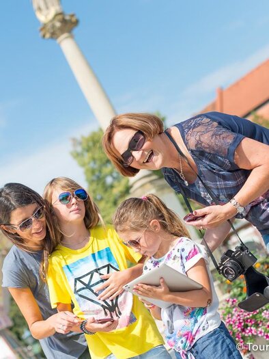 Eine Gruppe von Frauen und Kindern genießt einen sonnigen Tag im Freien. Sie schauen zusammen auf ein Handy und lachen, umgeben von Blumen und einem Vergnügungspark. | © Tourismusverband Thermen- Vulkanland