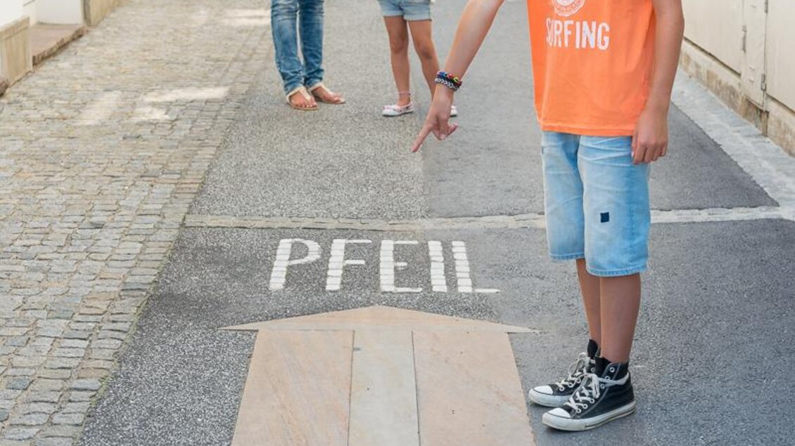 Eine fröhliche Familie steht auf einer Straße und zeigt auf ein großes Pfeil-Symbol auf dem Boden. Die Kinder tragen bunte T-Shirts und scheinen Spaß zu haben. | © Tourismusverband Thermen-Vulkanland