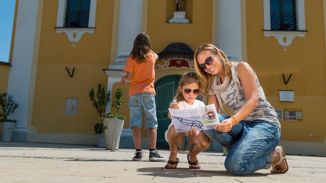Eine Mutter kniet mit ihrer Tochter auf dem Platz und schaut auf eine Karte. Im Hintergrund steht ein Junge und die historische Architektur ist sichtbar. | © Tourismusverband Thermen-Vulkanland