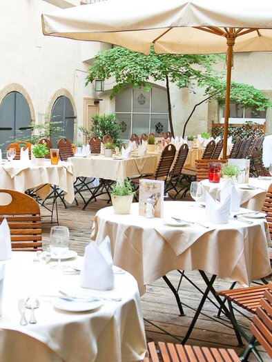 An inviting outdoor area of a restaurant with wooden tables and chairs. The space is decorated with tablecloths and plants, ideal for a relaxed meal al fresco. | © Stainzerbauer