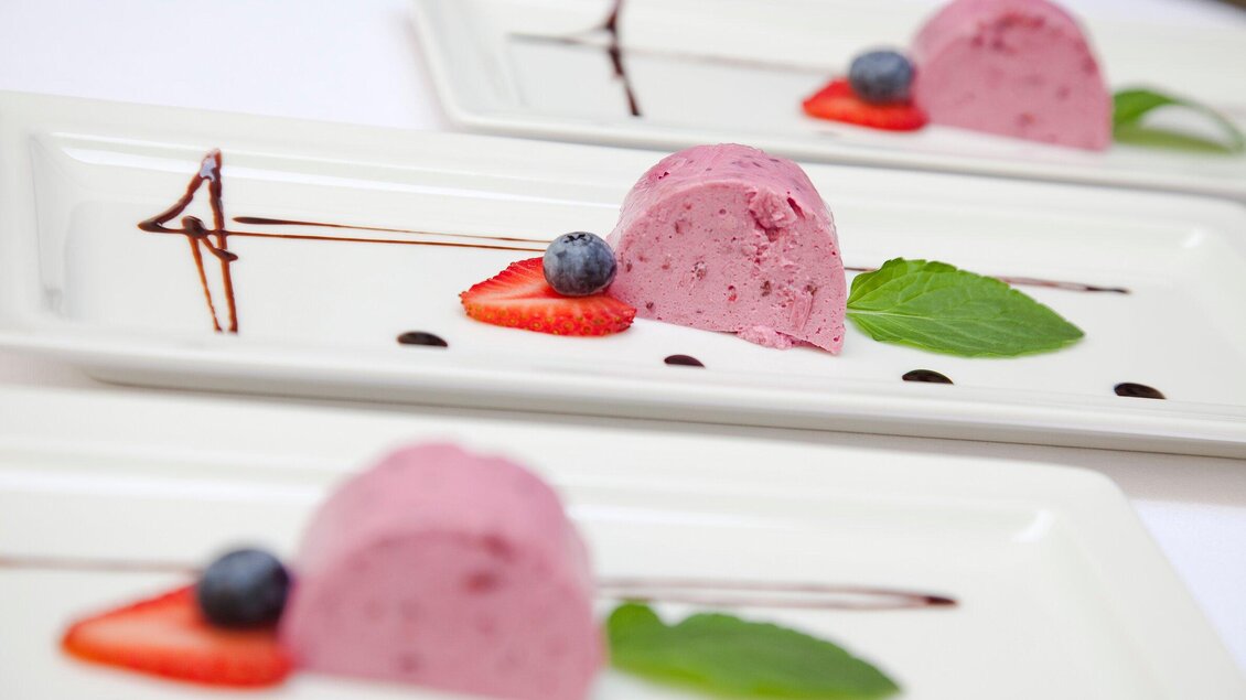Ein eleganter Dessertteller mit einer halbrunden Himbeermousse, garniert mit frischen Erdbeeren, Heidelbeeren und Minzblättern. Kunstvolle Schokoladensauce verleiht dem Gericht eine gehobene Note. | © Stainzerbauer