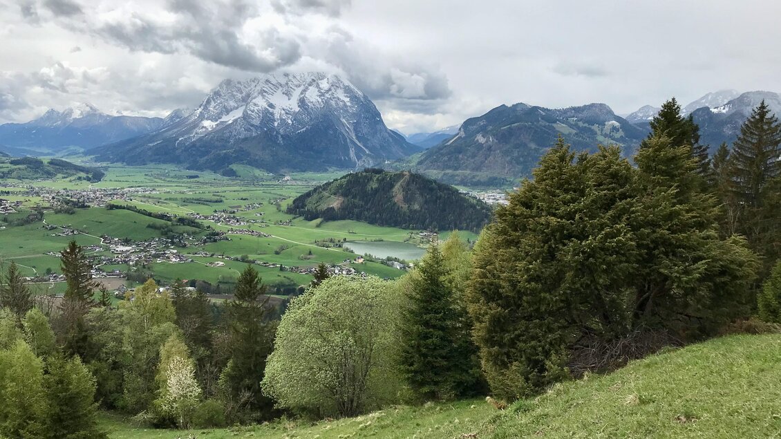 Eine malerische bergige Landschaft mit grünen Wiesen und schneebedeckten Gipfeln. Im Hintergrund ist ein ruhiges Tal mit einem kleinen Ort zu sehen. | © Sabine Schulz