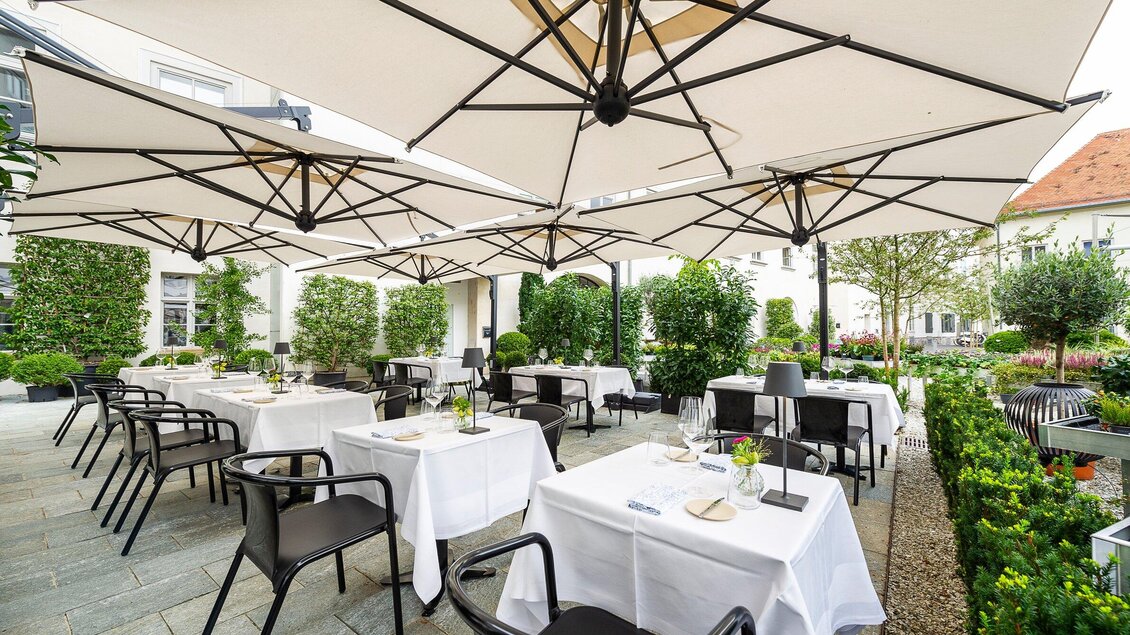 Ein elegantes Restaurant mit Tischen im Freien, die mit weißen Tischtüchern gedeckt sind. Große Sonnenschirme bieten Schatten und üppiges Grün umrahmt den Bereich. | © Stammtisch - Paul Stajan