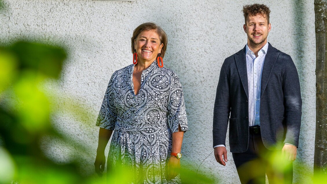 Ein lächelndes Paar steht vor einer grauen Wand. Die Frau trägt ein gemustertes Kleid und große Ohrringe, der Mann ist in einem dunkelblauen Blazer gekleidet. | © Stammtisch am Paulustor