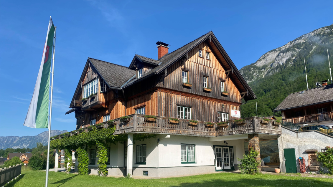 Ein traditionelles Holzhaus mit einem großen Balkon und Blick auf die Landschaft. Im Hintergrund sind Berge und ein blauer Himmel sichtbar. | © Bettina Scheck