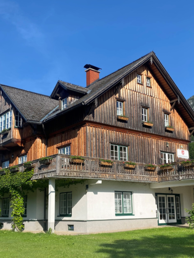Ein traditionelles Holzhaus mit einem großen Balkon und Blick auf die Landschaft. Im Hintergrund sind Berge und ein blauer Himmel sichtbar. | © Bettina Scheck