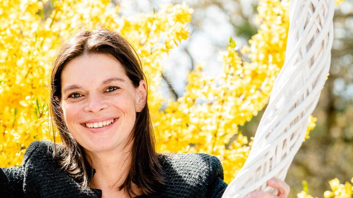 Eine lächelnde Frau steht vor einem Hintergrund aus gelben Blüten. Sie trägt eine schwarze Jacke und wirkt fröhlich. | © Region Graz - Mias Photoart