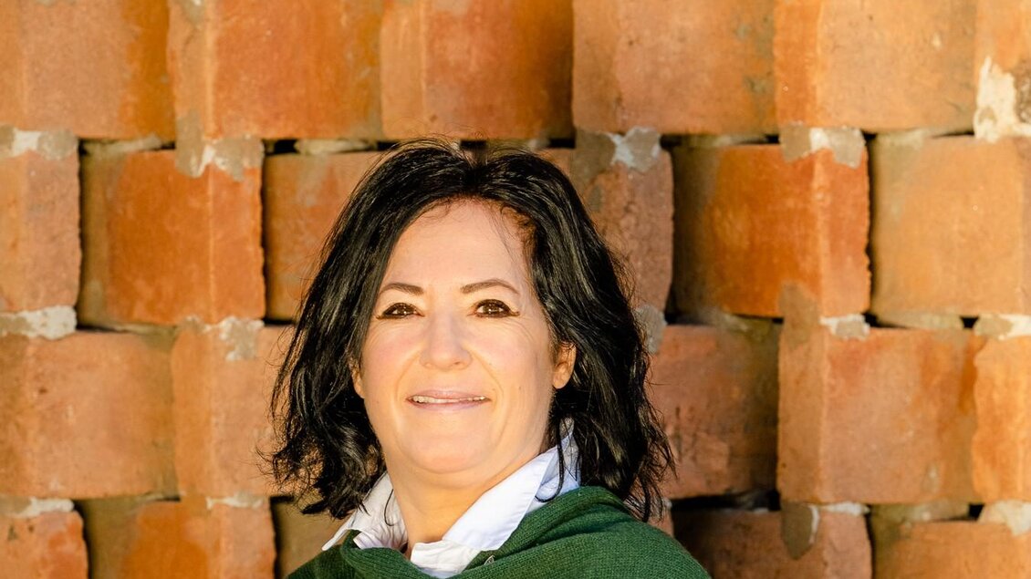 Eine Frau mit einem grünen Schal steht vor einer Wand aus Ziegelsteinen. Sie lächelt freundlich in die Kamera.