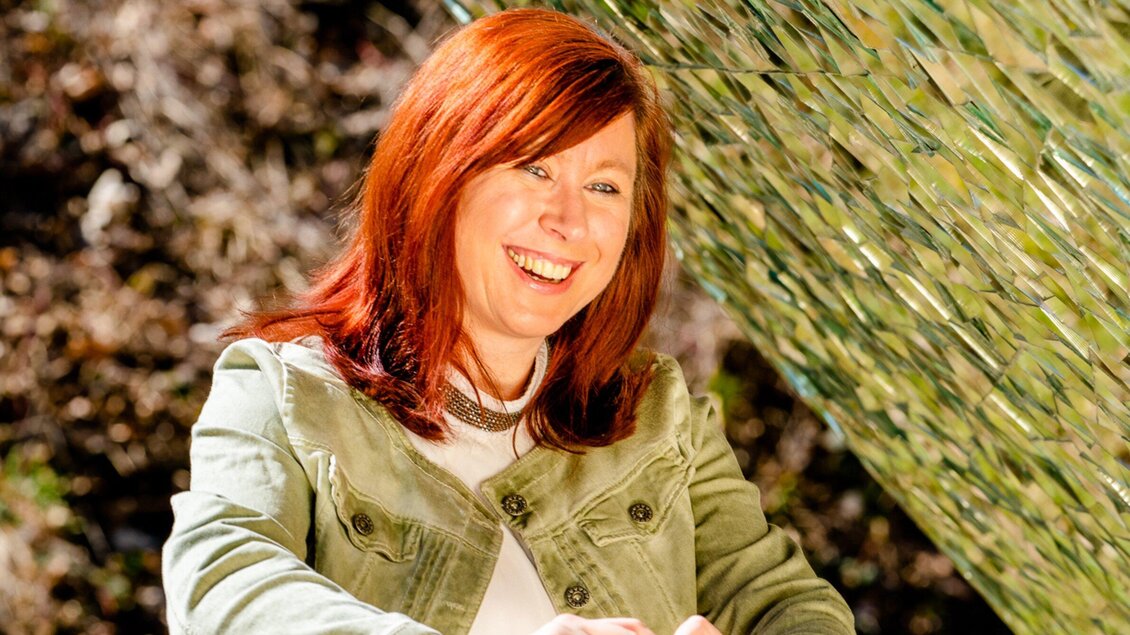Eine lächelnde Frau mit rotem Haar sitzt vor einem grünen, schilfartigen Hintergrund. Sie trägt eine Jacke und wirkt entspannt und fröhlich. | © TV Region Graz - Mias Photoart