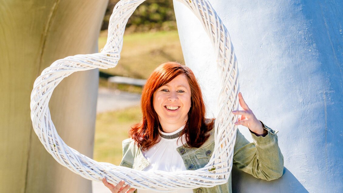 Eine lächelnde Frau steht hinter einem herzförmigen Rahmen. Sie trägt eine Jacke und hält den Rahmen lächelnd in den Händen. | © TV Region Graz - Mias Photoart