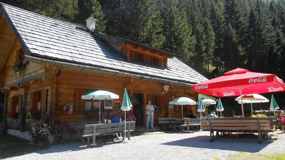 Eine gemütliche Berghütte umgeben von Bäumen. Draußen stehen Tische mit Sonnenschirmen für Gäste. | © Stangl Alm