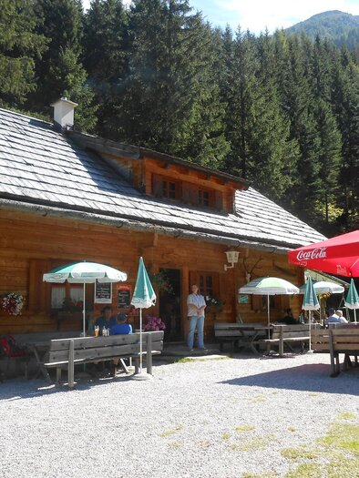 A cozy mountain cabin surrounded by trees. Outside, there are tables with umbrellas for guests. | © Stangl Alm