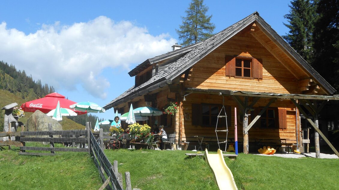 Eine gemütliche Holzhütte umgeben von grünen Wiesen und Bäumen. Im Vordergrund gibt es eine Rutsche und Sonnenschirme zum Entspannen. | © Stangl Alm