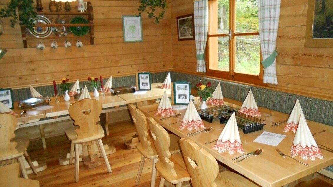 Ein gemütlicher Raum mit Holzwänden und einem großen Tisch, der für eine Feier dekoriert ist. Es gibt gefaltete Servietten und Blumenschmuck auf jedem Tisch. | © Stangl Alm