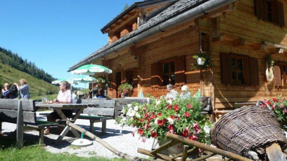 Ein gemütliches Holzhaus in den Bergen mit blühenden Blumen. Menschen genießen die frische Luft und die Sonne auf einer Terrasse. | © Stangl Alm