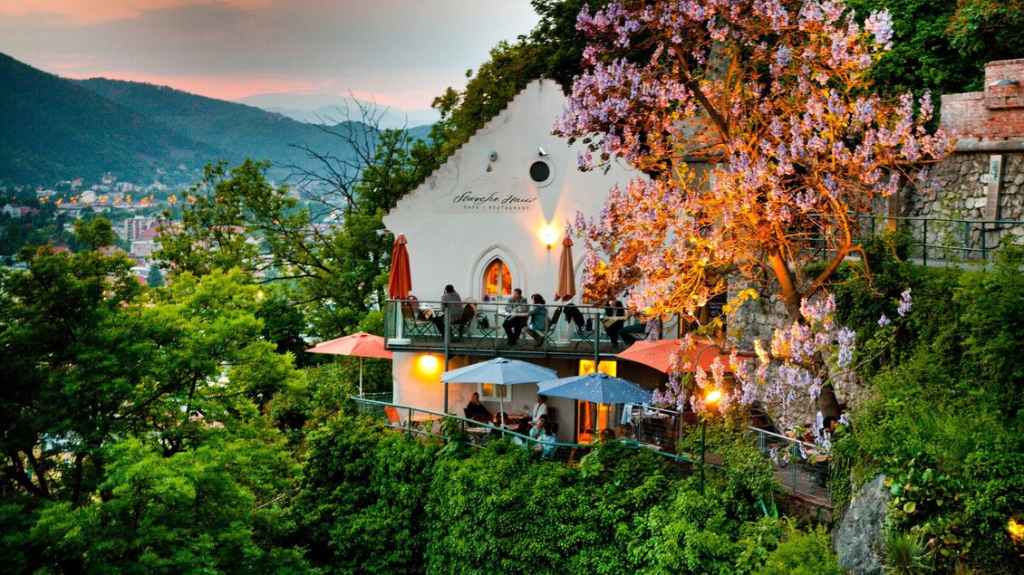 Ein charmantes Restaurant auf einer Terrasse, umgeben von Bäumen und blühenden Pflanzen. Im Hintergrund sind sanfte Hügel und der Abendhimmel zu sehen. | © Graz Tourismus - Werner Krug