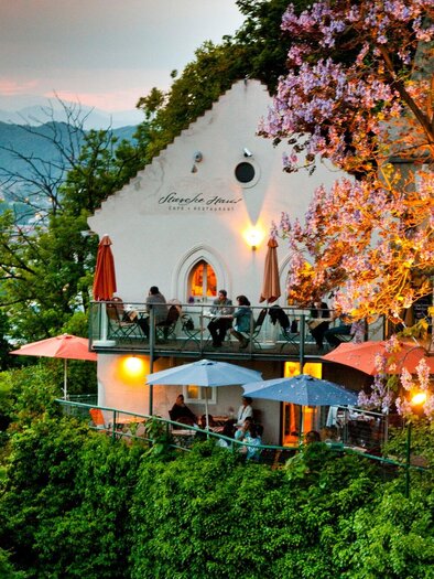 A charming restaurant on a terrace, surrounded by trees and blooming plants. In the background, gentle hills and the evening sky can be seen. | © Graz Tourismus - Werner Krug