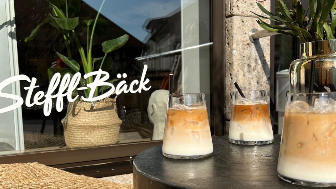 Drei Gläser mit Eiskaffee stehen auf einem Tisch vor einem Fenster. Im Hintergrund sind Pflanzen und das Logo „Steffl Bäck“ sichtbar. | © Bäckerei Steffl-Bäck
