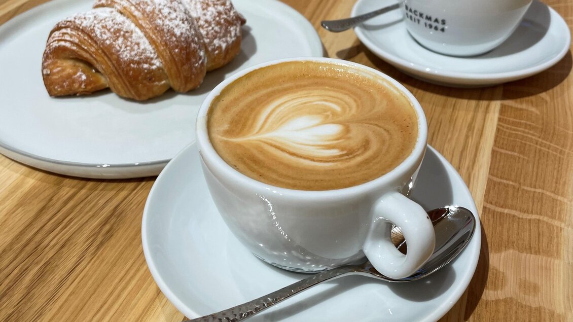 Eine Tasse Kaffee mit kunstvollem Milchschaum und ein Croissant auf einem Teller. Im Hintergrund ist eine zweite Tasse Kaffee zu sehen. | © Steffl-Bäck Sebastian Knapp